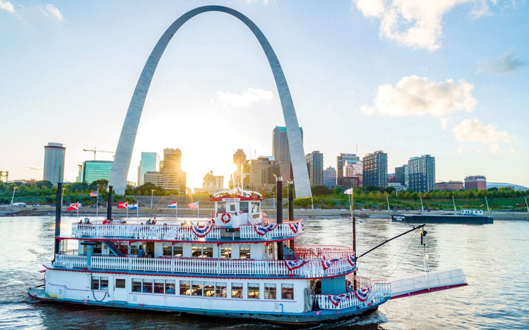 A white riverboat sails past the St. Louis skyline, which is dominated by the sight of the soaring Gateway Arch.