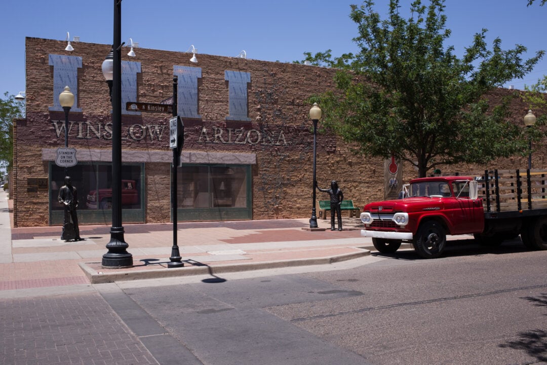 a brick building on a corner with two bronze statues and a red flatbed ford truck parked nearby, winslow arizona