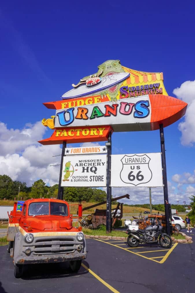 A colorful sign for the Uranus Fudge Factory with a motorcycle and an old truck parked in front of it