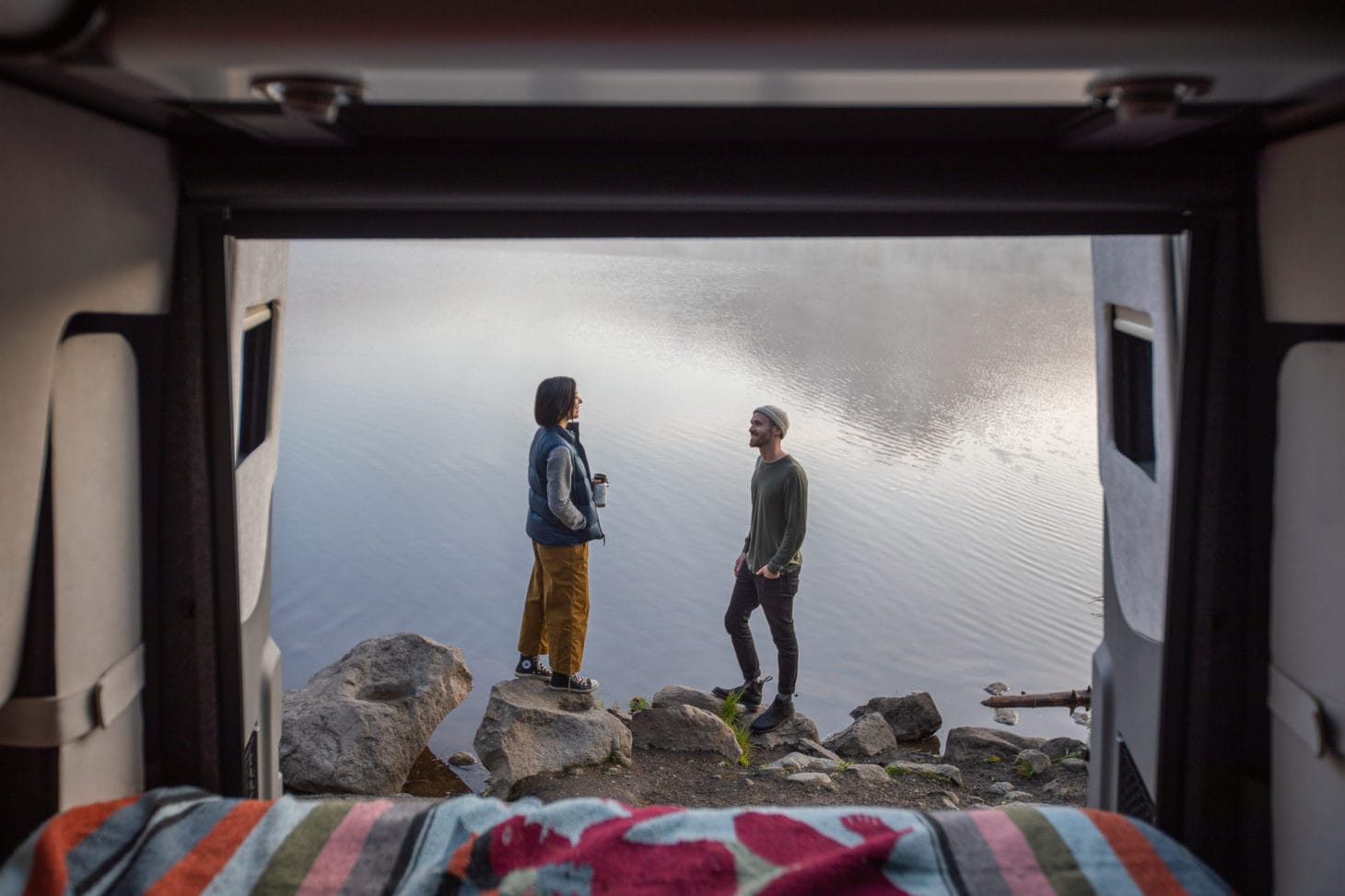 Van backed out onto water view with two people standing on rocks.