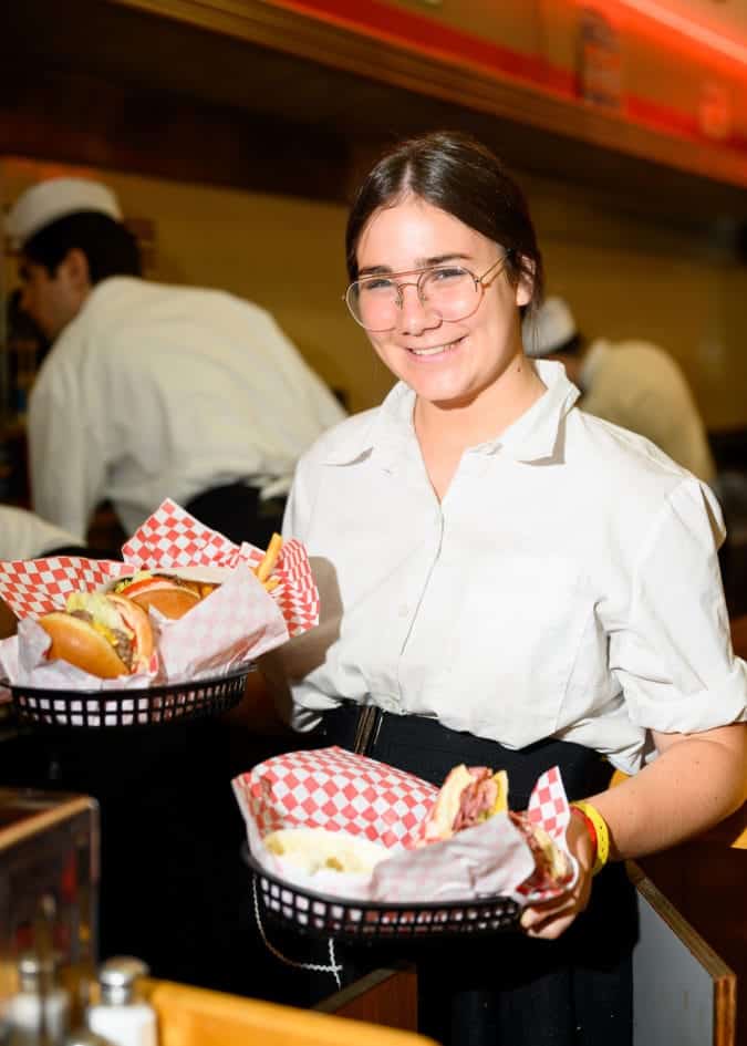 a young girl in glasses holds to baskets filled with burgers at woolworth's diner