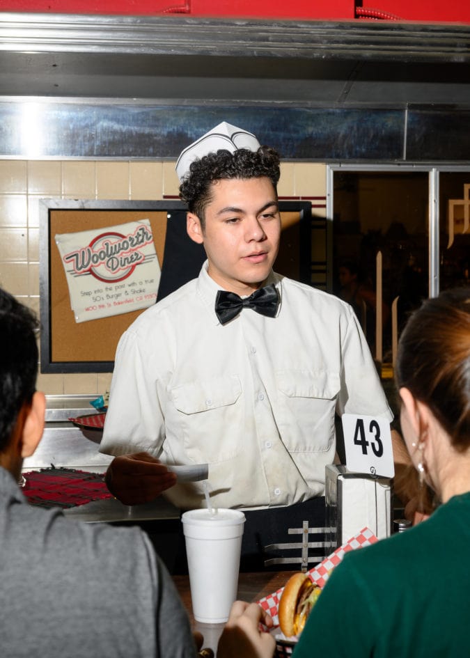 a man in a button up shirt, bowtie, and hat helps customers at woolworth's diner